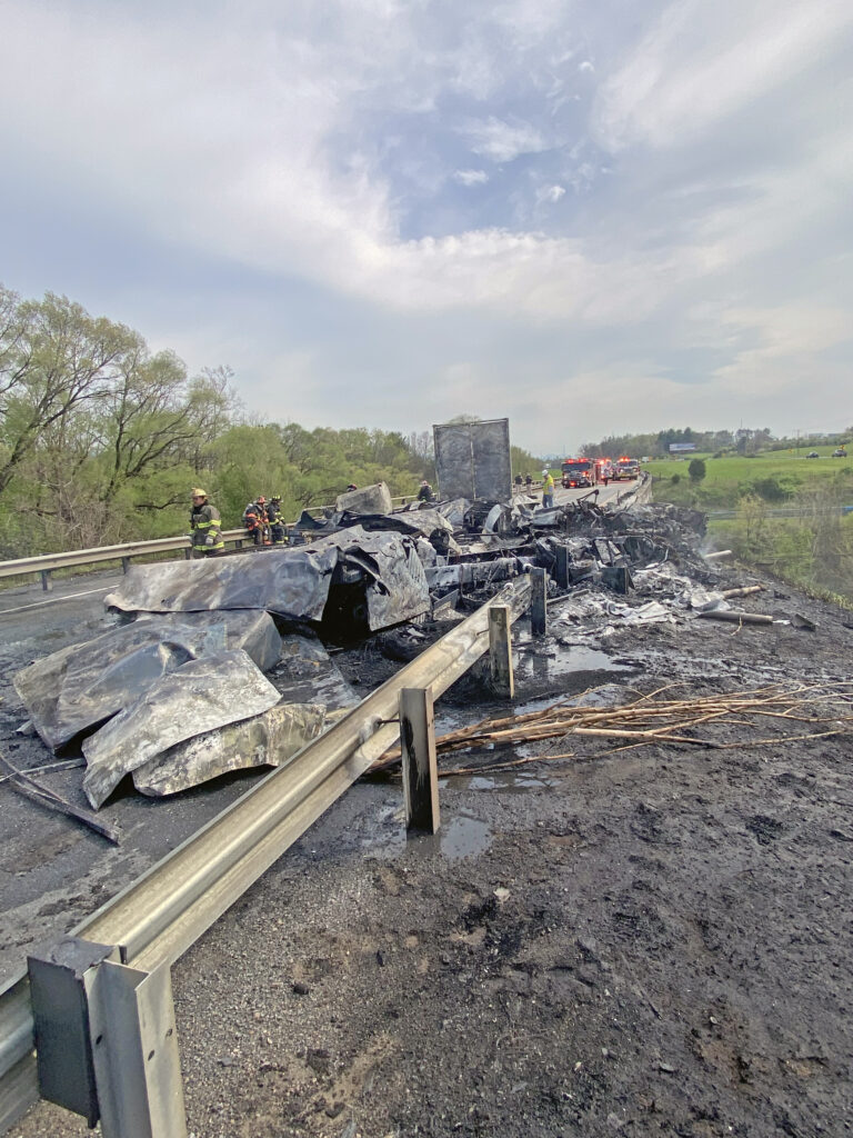 Charred remains of a tractor-trailer on the shoulder of Interstate 81 after a fire, with emergency vehicles and personnel on scene managing the aftermath.